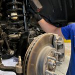 Close-up of a car's exposed wheel assembly showing the brake rotor, caliper, coil spring, and a mechanic working on the shock absorber.